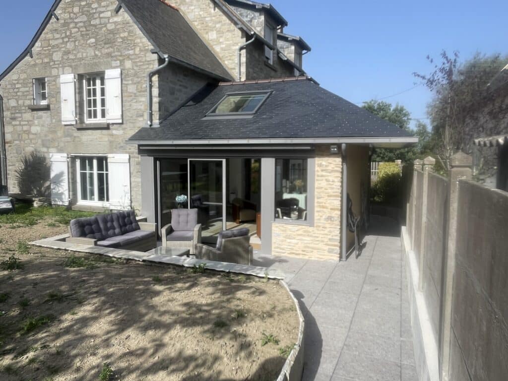 Extension de Maison Moderne en Pierre et Verre avec Patio Extension moderne à toit en ardoise et baies vitrées sur maison traditionnelle en pierre avec salon de jardin.