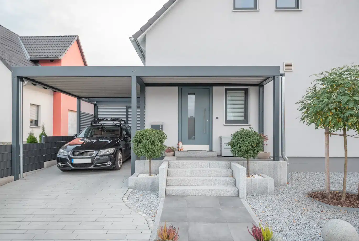 Carport gris anthracite attaché à une façade moderne. La zone d'entrée est aménagée avec des escaliers en granit, une porte bleu-gris et une allée pavée.