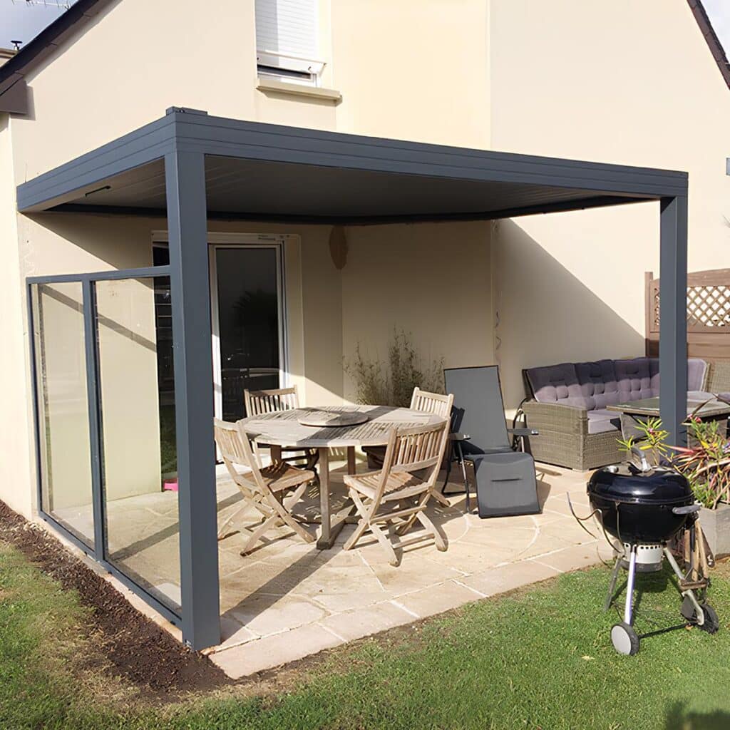 Aménagement de terrasse moderne avec pergola anthracite. Pergola anthracite moderne sur une terrasse aménagée. Mobilier de jardin en bois, salon lounge en rotin et cloisons vitrées contre le vent.