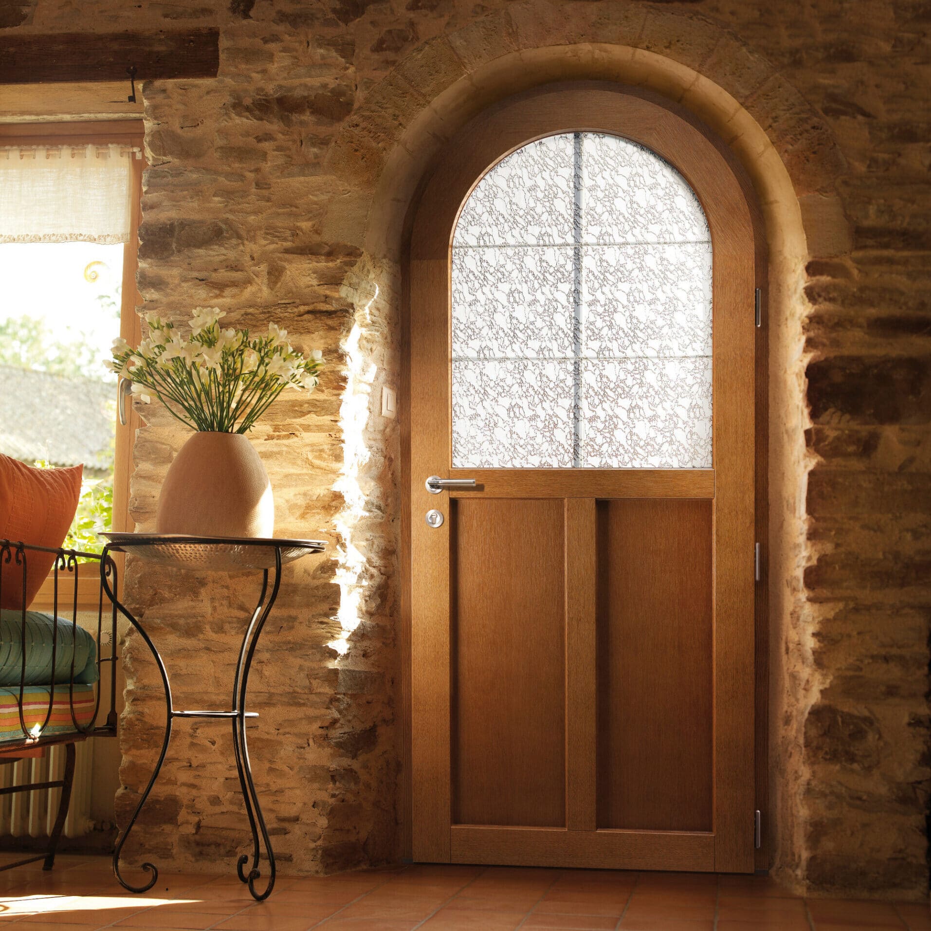 Porte en bois cintrée avec vitrage décoratif intégrée dans une épaisse arche de pierre rustique, éclairée par le soleil.