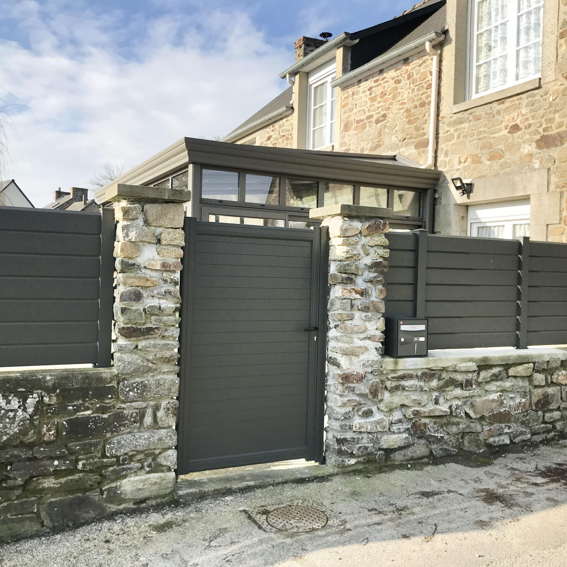 Portillon et clôture en aluminium gris anthracite à lames devant des piliers et un mur en pierres. Maison ancienne en arrière-plan.