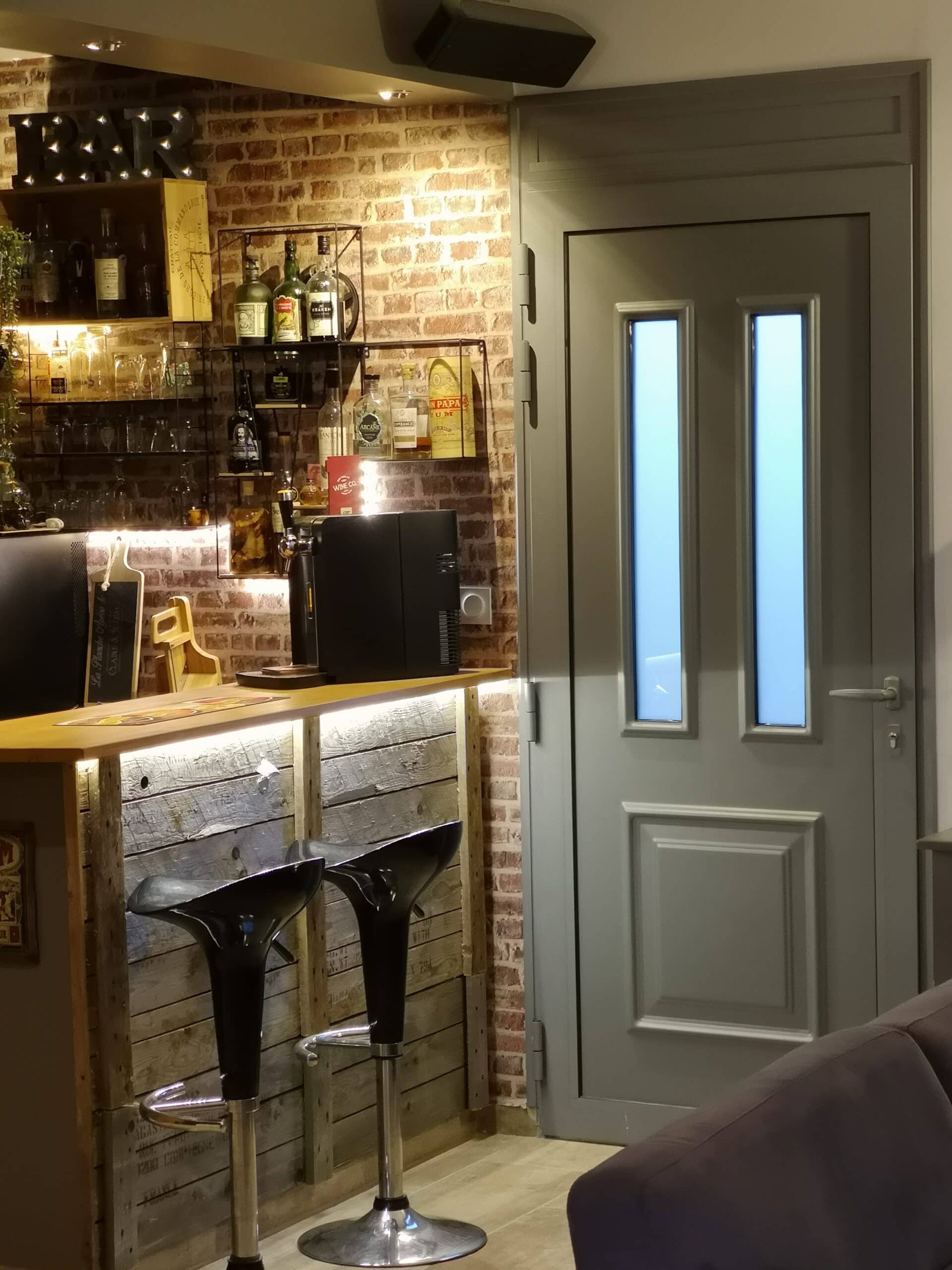 Bar rustique chic : mur de briques, tabourets noirs design, étagères d'alcools et porte grise avec vitres dépolies.