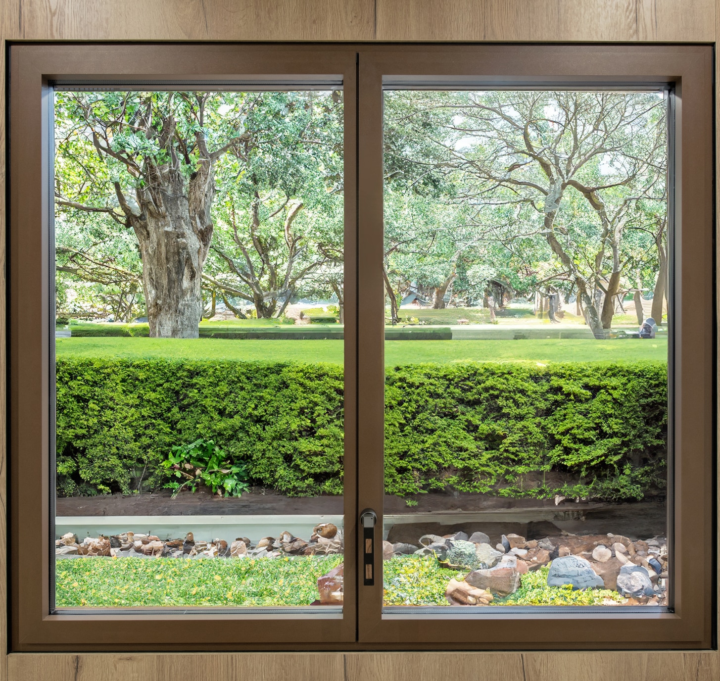 Fenêtre à cadre marron avec vue sur un jardin verdoyant, grand arbre, haie taillée et rocailles décoratives.