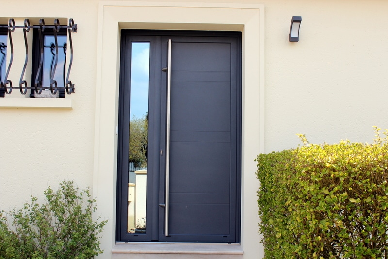 Porte d'entrée moderne gris anthracite avec vitrage latéral et longue poignée en inox sur façade beige.