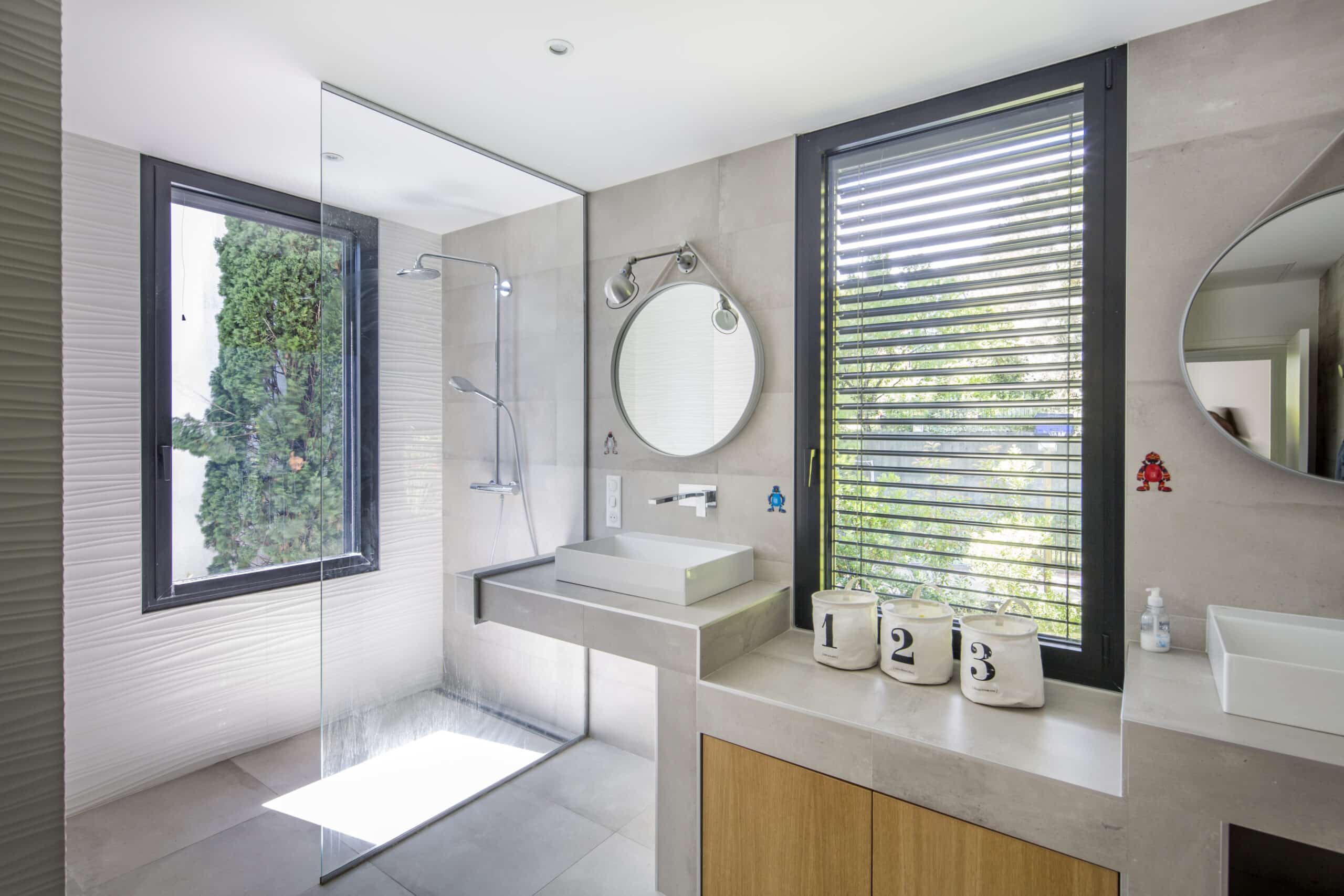 Salle de bain moderne lumineuse avec douche vitrée, vasque blanche, miroirs ronds et vue sur jardin extérieur.