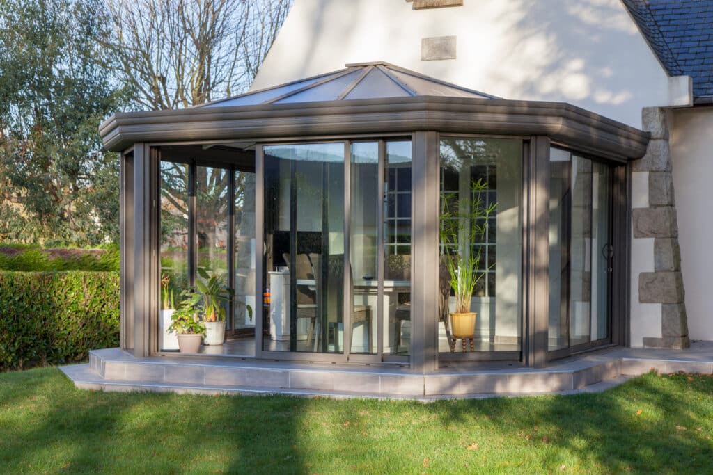 Véranda moderne en aluminium pour extension de maison Véranda moderne en aluminium gris avec grandes baies vitrées coulissantes sur terrasse carrelée et jardin verdoyant.
